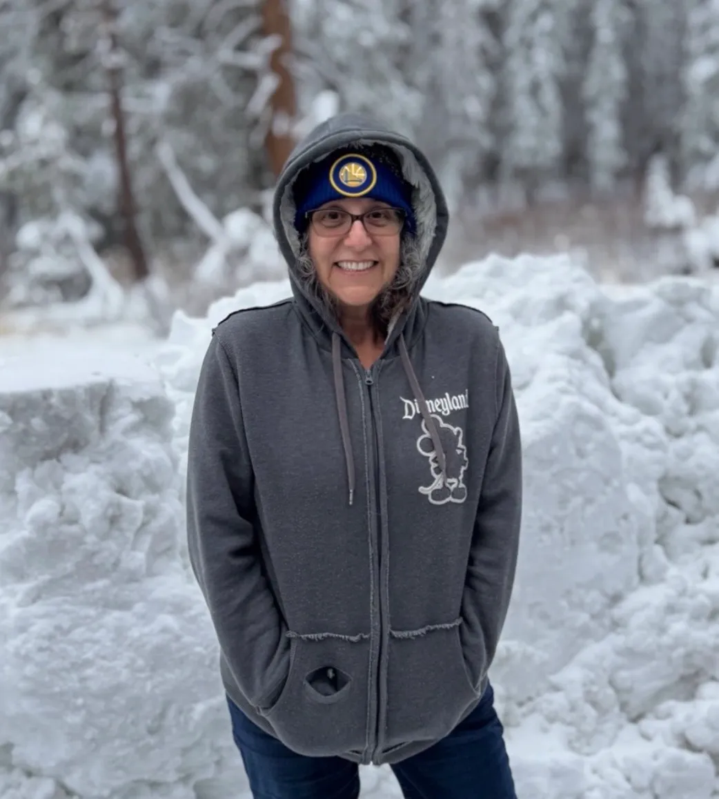 Corrinne Goldberg smiling in a hooded sweatshirt and beanie in a snowy forest.